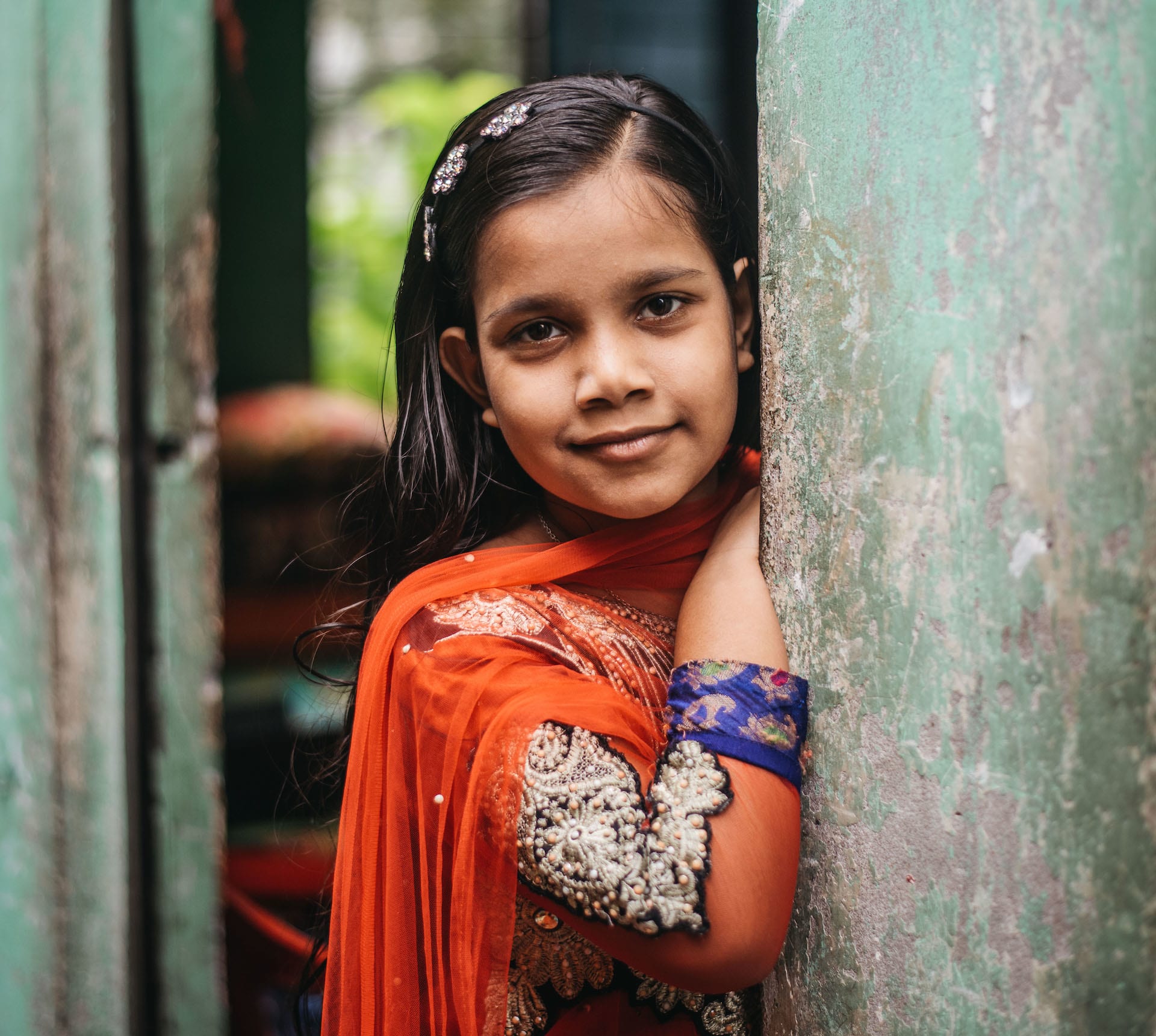 Mim leans against a green wall wearing an orange dress and a flower headband.